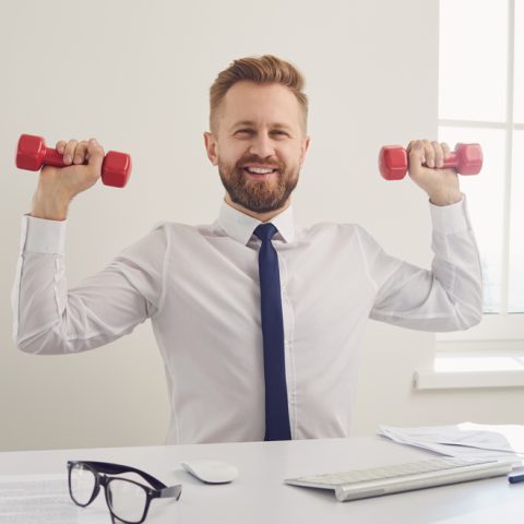 Eine männliche Person sitzt an einem Schreibtisch und hat Hanteln in der Hand