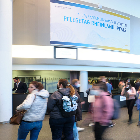 Foto aus der Veranstaltungshalle mit mehreren Personen. Text auf einem Wanddisply: Pflegetag Rheinland-Pfalz